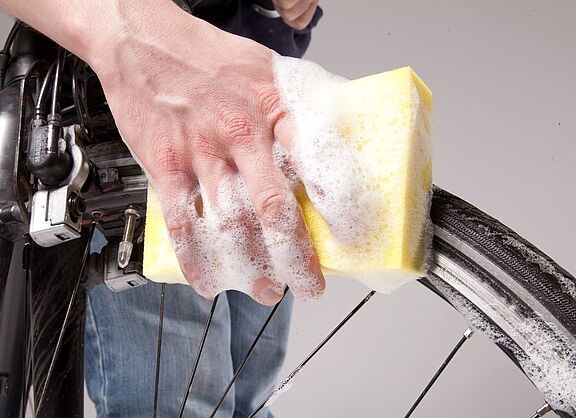 Großaufnahme einer Hand mit Schwamm und Schaum, die ein Fahrrad-Vorderrad säubert. 