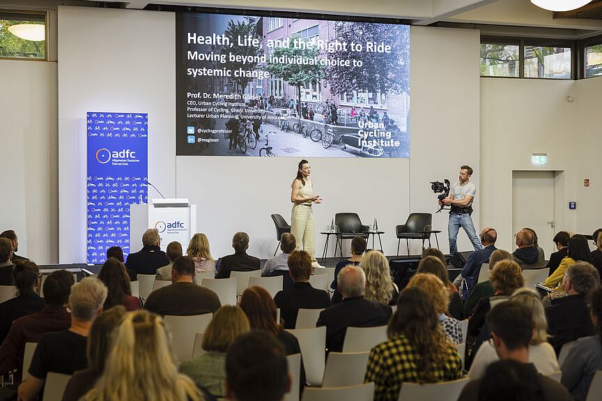 Auf dem Internationalen ADFC-Symposium hielt Prof. Dr. Meredith Glaser vom Urban Cycling Institute eine Keynote. Internationales ADFC-Symposium "Gesundheit x Mobilität": Sprecherin mit Publikum und Kameramann
